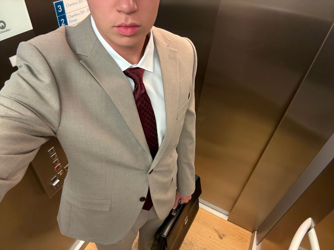 Light stone single-breasted suit with notch lapels and flap pockets, worn with a white shirt and a burgundy tie.